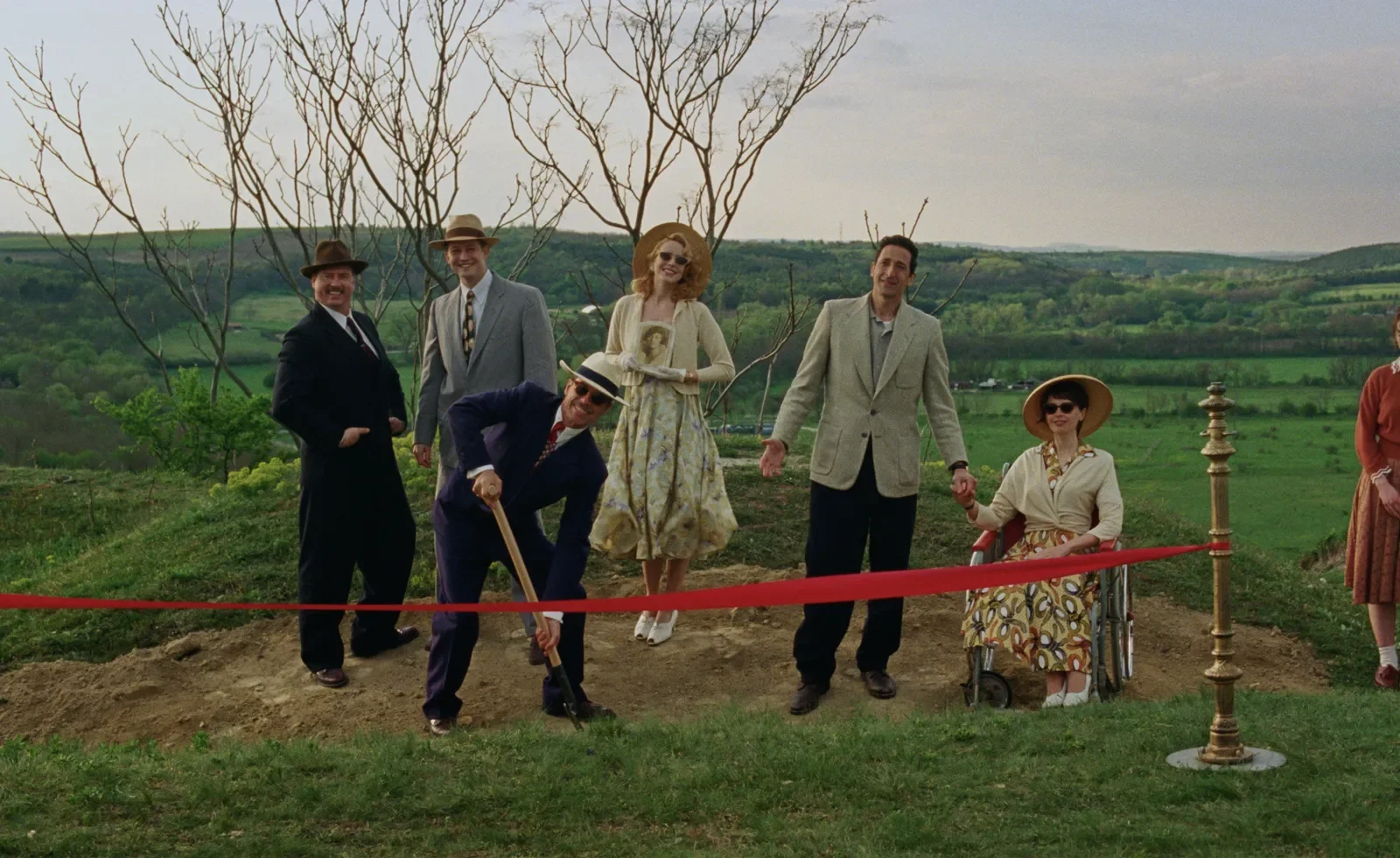 A group of six people in elegant 1950s attire takes part in a groundbreaking ceremony on a lush green hill. A man in a dark suit and hat digs with a shovel, while a woman in a wheelchair and other guests smile. The background reveals rolling countryside under a slightly cloudy sky.