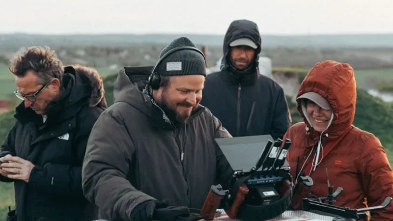 A warmly dressed film crew works outdoors on the set of The Brutalist. At the center, a smiling director checks a monitor, surrounded by technicians and a camera operator equipped with a stabilizing rig. The background shows a rural landscape under an overcast sky.