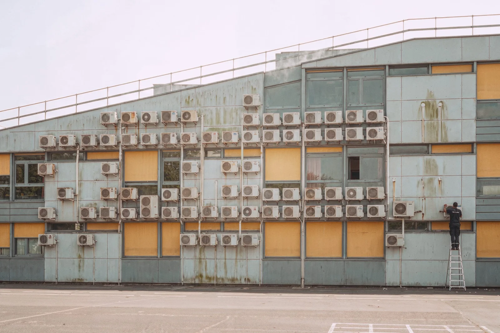 Une façade d'école couverte d'une centaine de climatiseurs extérieurs répartis sur trois niveaux, un technicien en installant un de plus au sommet d'une échelle.