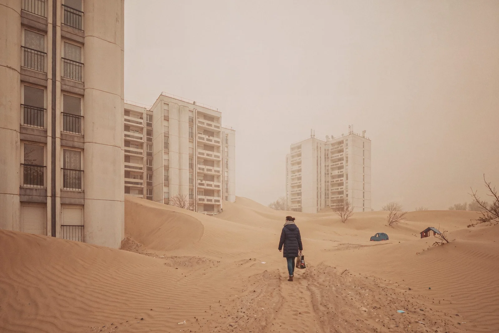 Un cœur d'îlot de grands ensembles enseveli sous des dunes de sable qui atteignent les rez-de-chaussée, une aire de jeux à demi enterrée, une femme marchant avec un cabas.