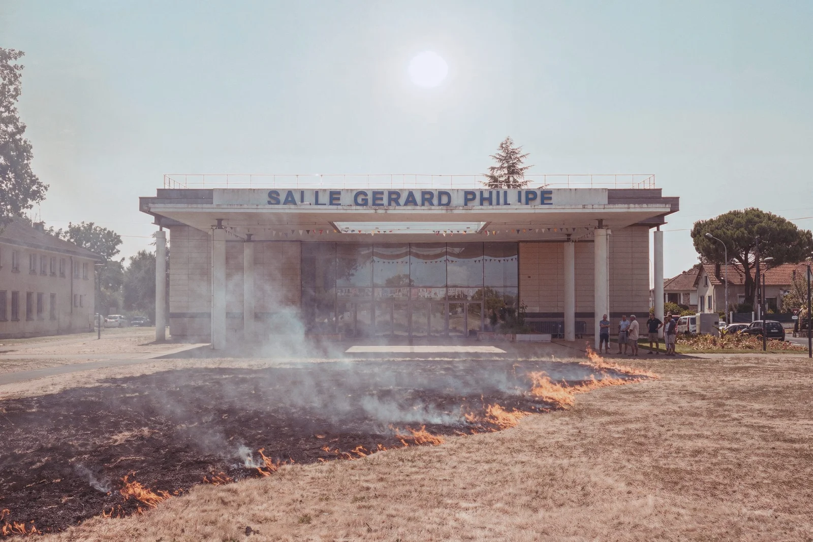 Une ligne de flamme basse avançant sur une pelouse calcinée devant une salle municipale, cinq personnes immobiles sur le parvis, le soleil pâli par la fumée.